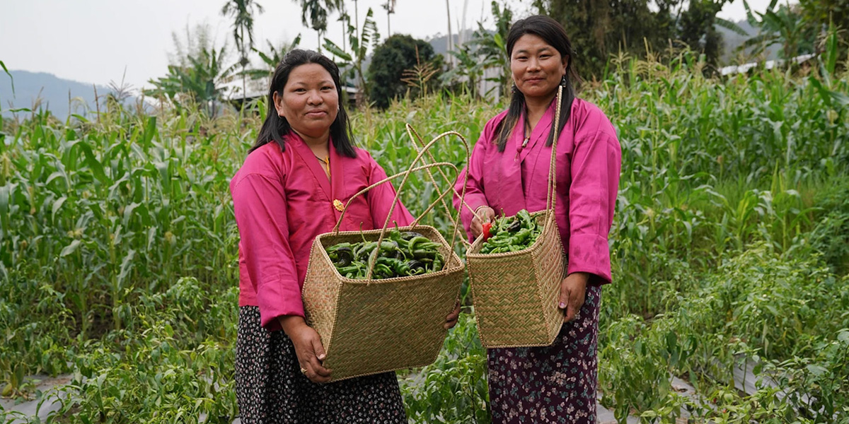 Two women farmers in Bhutan stand in a green field, smiling and holding baskets filled with freshly harvested green chilies.