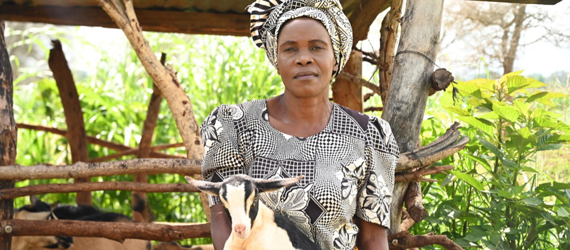 Zambian woman farmer in traditional dress holding a goat by a domestic pen.