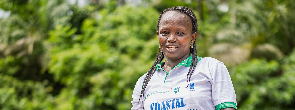 Woman standing outdoors in Guinea-Bissau wearing a coastal resilience programme shirt, with tropical vegetation in the background.