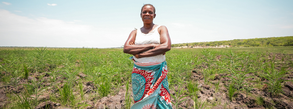 Woman farmer standing in a cultivated field, arms crossed, surrounded by young crops in a rural landscape.