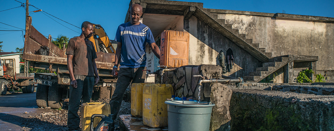 Two men collect water in plastic containers from a public tap in Moroni, Comoros, with jerry cans and buckets lined up beside a concrete water station.