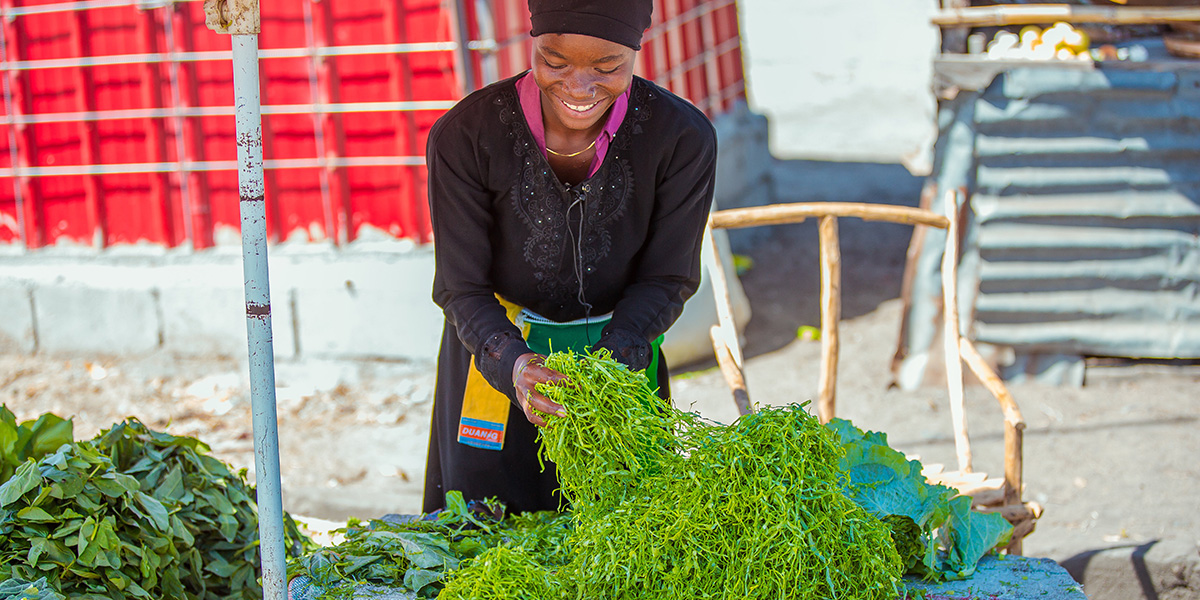 A woman vendor smiles while arranging fresh green vegetables at a market stall in Mozambique.
