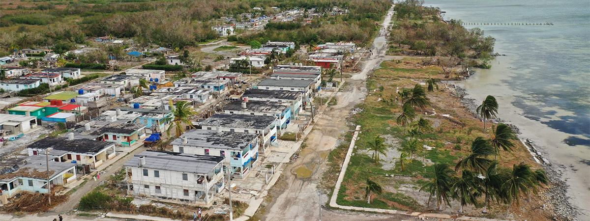 Aerial view of a coastal town with damaged homes, flooded streets, and uprooted palm trees along the shoreline after a severe storm.