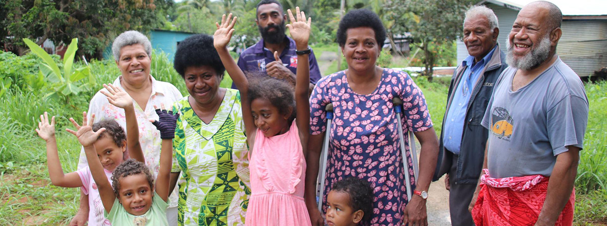 Community members of different generations in Fiji smile and wave together outdoors in a village, including children, adults and an older person.