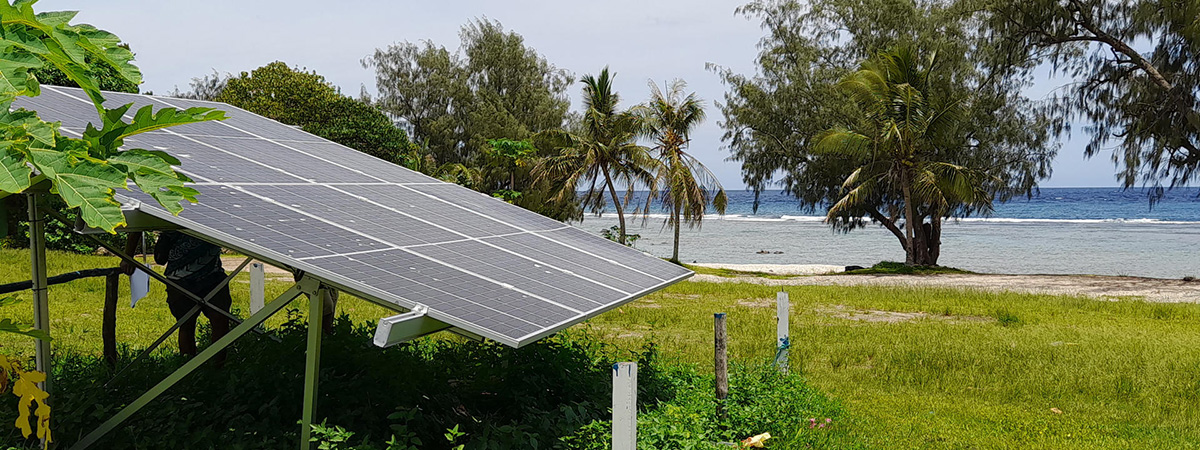 Solar panels stand on a grassy shoreline surrounded by palm trees and vegetation overlooking the ocean in Vanuatu.