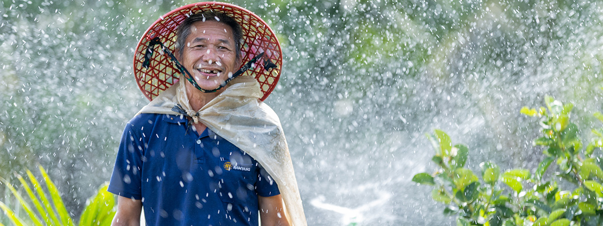 A smiling farmer wearing a traditional woven hat stands in an irrigated field as water sprays over surrounding crops.