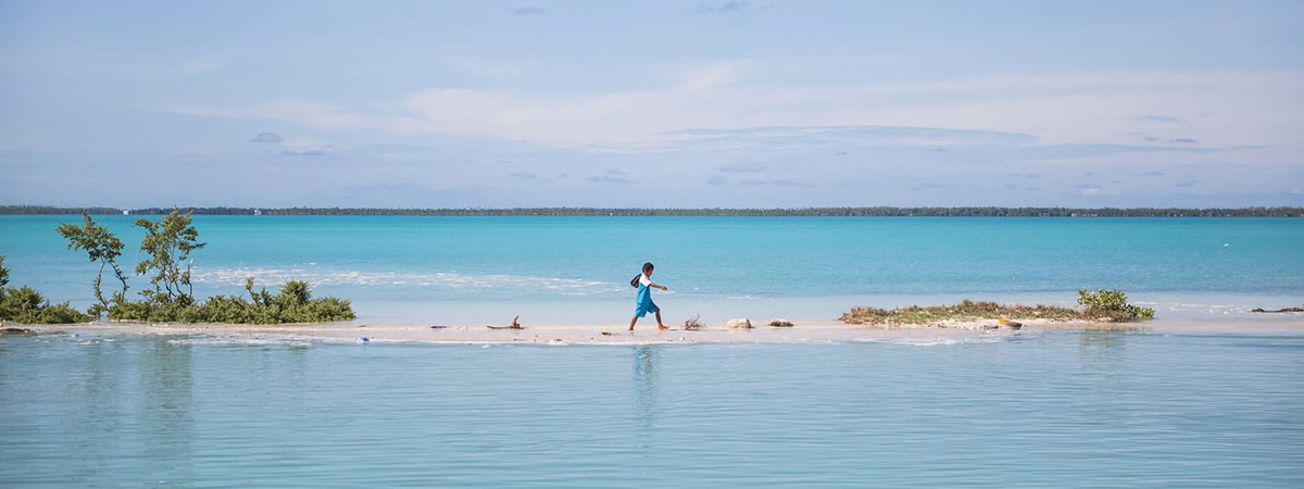 A child walks along a narrow sandbar surrounded by turquoise lagoon waters in a low-lying Pacific atoll.