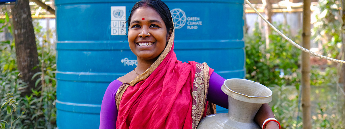A smiling woman in traditional Bangladeshi clothing stands beside a household rainwater storage tank, holding a water container.