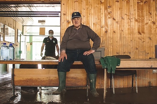 A man in rubber boots takes a break from cleaning, sitting on a desk in a flood-damaged room, with muddy floors and water bottles