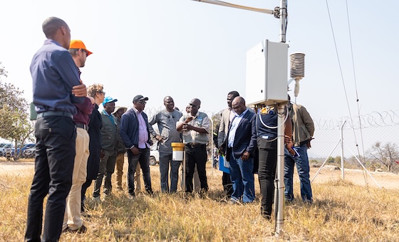 A group of people stands in a dry, grassy field, attentively listening to a presenter explaining the function of a weather monitoring station