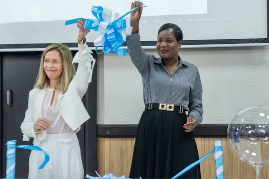 Two women stand at a table with blue and white balloons; one wears a white lab coat.